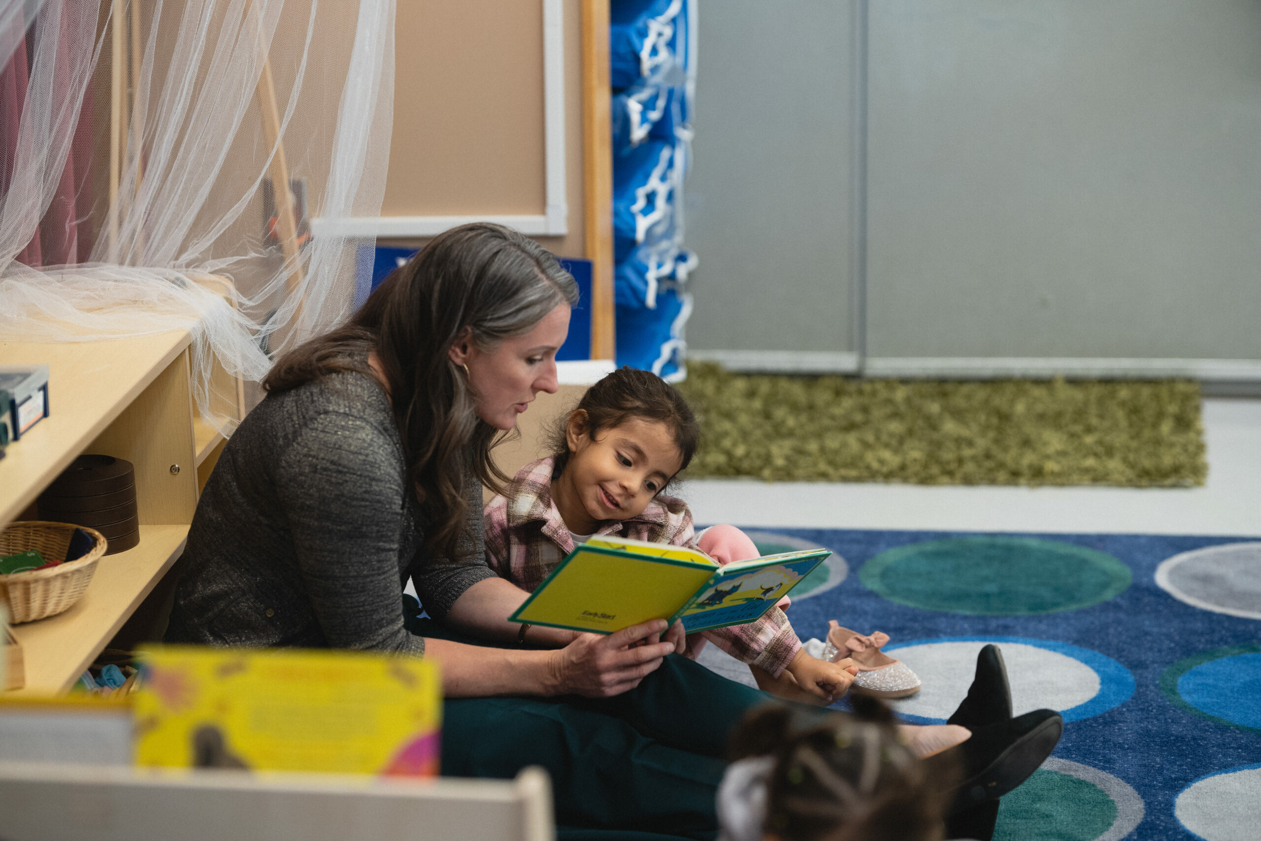 Adult reads smiling child a story on a rug in a classroom